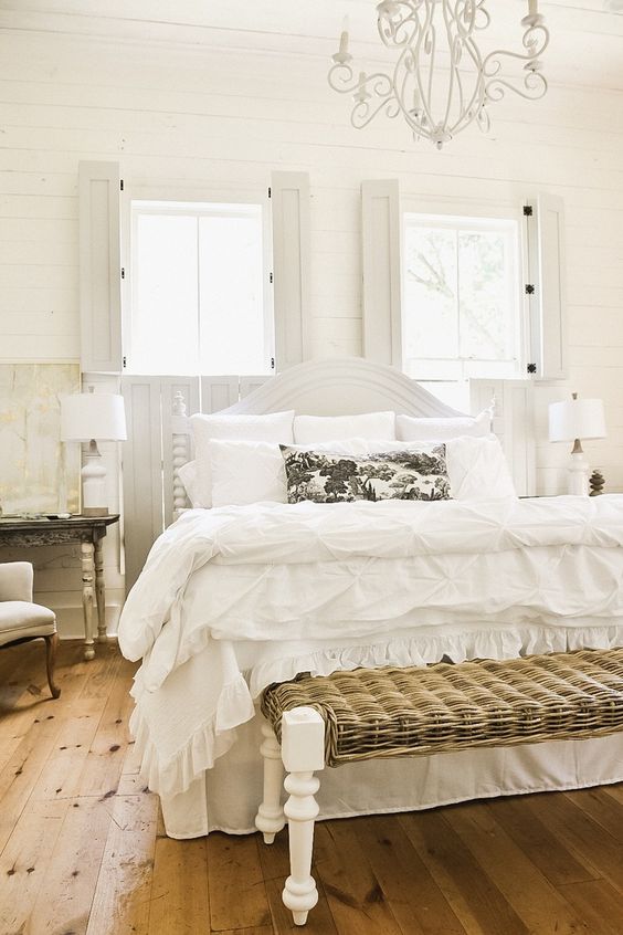 a neutral Provence bedroom with white vintage and shabby chic furniture, neutral textiles, neutral seating furniture, woodne shutters and a white chandelier