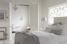 a neutral Provence bedroom with wooden beams, a window with a wrought cover, a creamy bed with white and grey bedding, artwork and a crystal chandelier