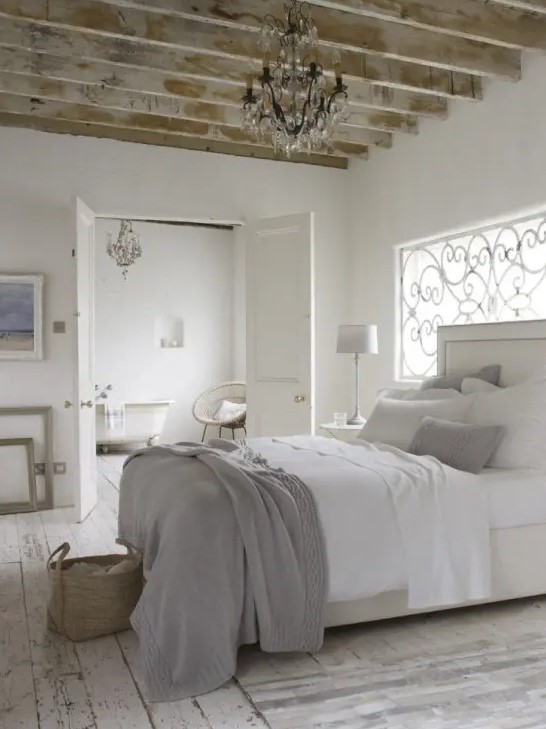 a neutral Provence bedroom with wooden beams, a window with a wrought cover, a creamy bed with white and grey bedding, artwork and a crystal chandelier