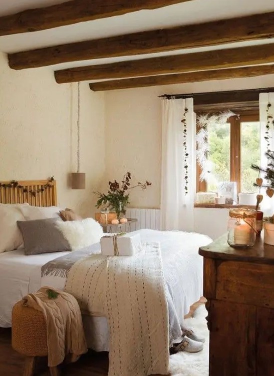 a neutral and cozy Provence bedroom with stained wooden beams, a bed with neutral bedding, a stained dresser, pendant lamps, candle lanterns and a jute pouf
