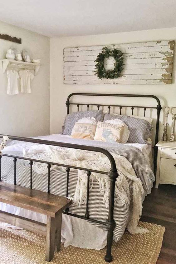 a neutral bedroom in vintage and shabby chic style, with a wooden artwork and a wreath, a forged bed, striped bedding and some neutral decor