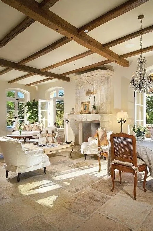 a neutral chic living room with wooden beams, a refined fireplace, neutral and sophisticated furniture, a crystal chandelier and lovely pillows