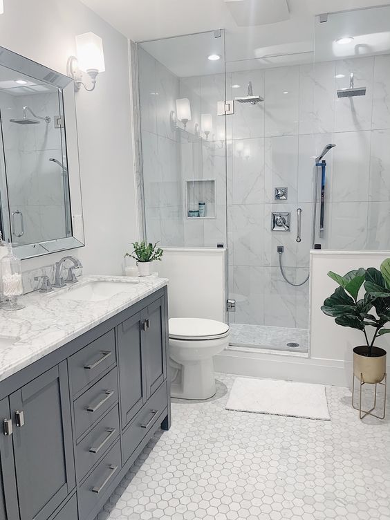 a neutral modern bathroom clad with marble hex tiles, with a shower space with half walls, a grey vnaity and a large mirror