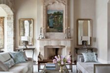 a neutral vintage living room with a fireplace, a refined chandelier, two mirrors, a couple of sofas and a glass table