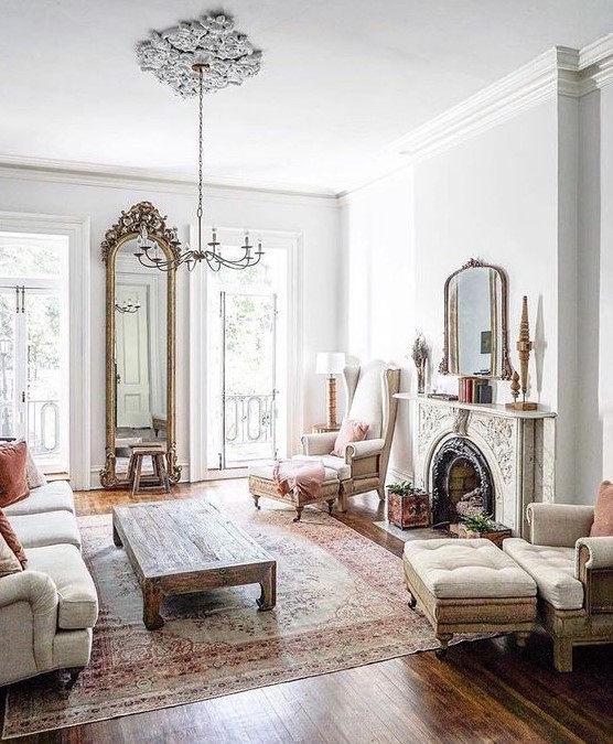 a neutral vintage living room with an ornate fireplace, neutral seating furniture, a low coffee table, a tall mirror in a fab frame and a chic chandelier
