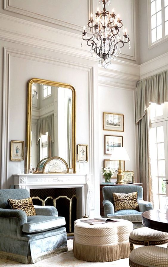 a neutral vintage living room with paneled walls, a fireplace, blue chairs, ottomans and stools, artworks and a large mirror