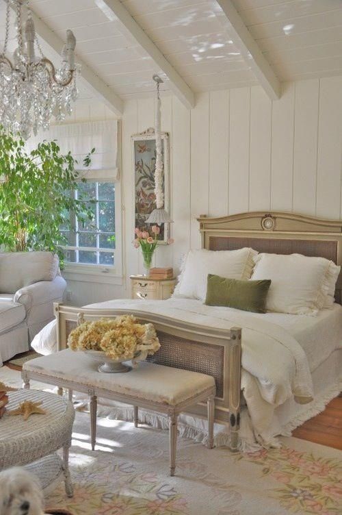 a refined French chic bedroom with shiplap, a refined bed with cane touches, lovely seating furniture, a crystal chandelier and some greenery