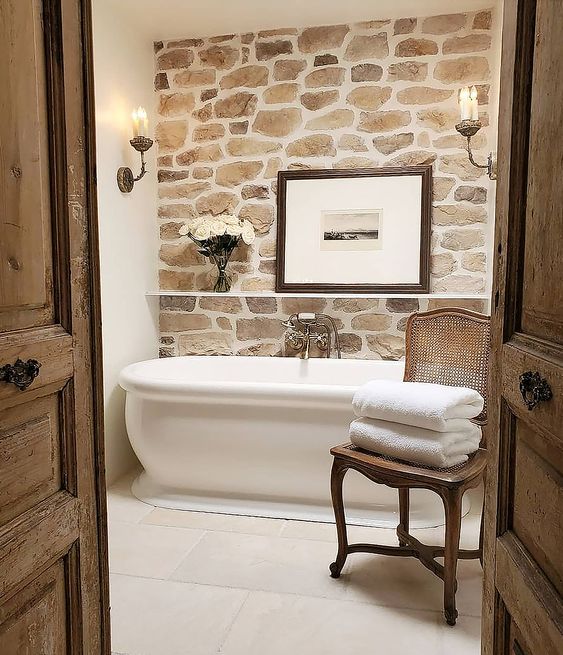 a small Provence style bathroom with a faux stone accent wall, a modern bathtub, wall lamps and blooms, a vintage chair