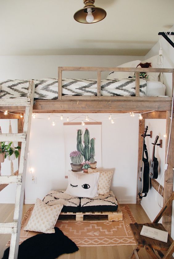 a small boho space with a pallet seat with pillows and a rug, guitars on the wall and string lights, a loft bed with a pendant bulb