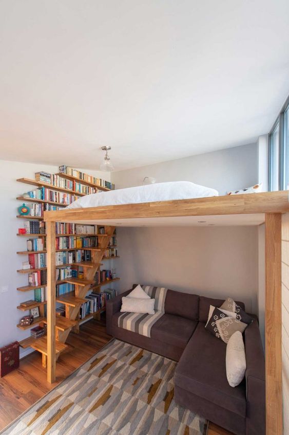 a small space with a corner sofa, a bookshelf that takes a whole wall and a loft bed with enough natural light