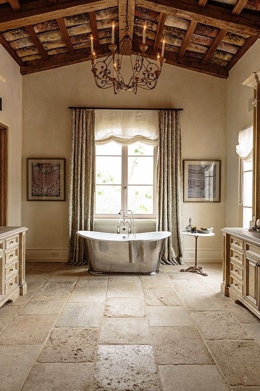 a soft buttermilk bathroom with textural stone floors and a wooden ceiling, a shiny metal bathtub, a beautiful metal chandelier and wooden vanity