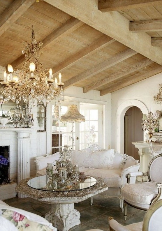 a sophisticated Provence living room with a fireplace, a crystal chandelier, a heavy table, neutral and refined furniture and wooden beams