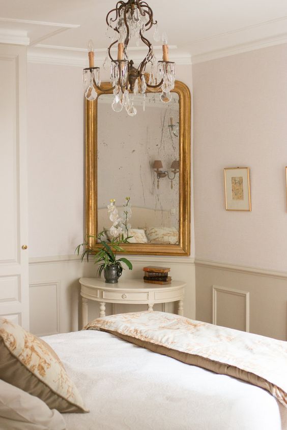 a sophisticated neutral bedroom with paneling, a vintage mirror, a crystal chandelier and chic bedding