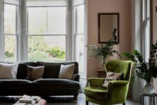 a stylish living room with pink walls, a grey sofa, a green chair, a pink one, a plaid ottoman, a printed rug and a chic chandelier