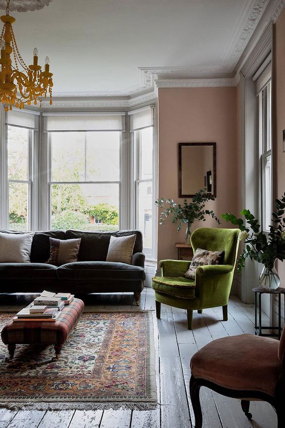 a stylish living room with pink walls, a grey sofa, a green chair, a pink one, a plaid ottoman, a printed rug and a chic chandelier