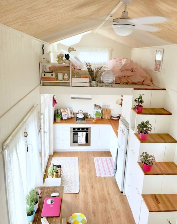 a tiny home with a loft bedroom, with some stands and storage units, a low bed with pastel bedding and some potted plants on the stairs