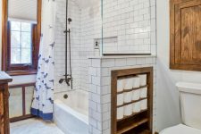 a vintage-inspired bathroom with white and marble tiles, with a half wall that has soem integrated storage space, touches of stained wood for more coziness