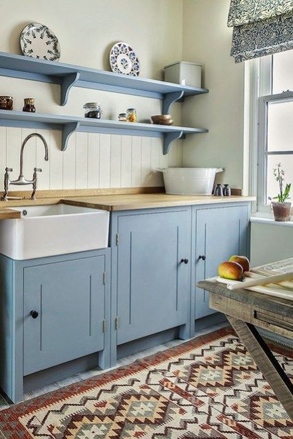 a vintage kitchen with blue cabinetry and shelves, a rough wooden table, a beadboard backsplash, a printed rug and butcherblock countertops