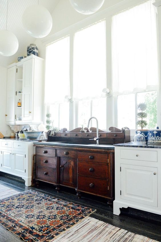 a vintage kitchen with white cabinets, a dark stained vanity of a vintage dresser, pendant lamps, printed rugs and lots of windows