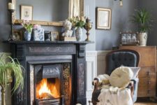 a vintage living roomw ith grey walls, a painted fireplace, a grey chair, a stained chest, greenery, a large mirror in a gilded frame