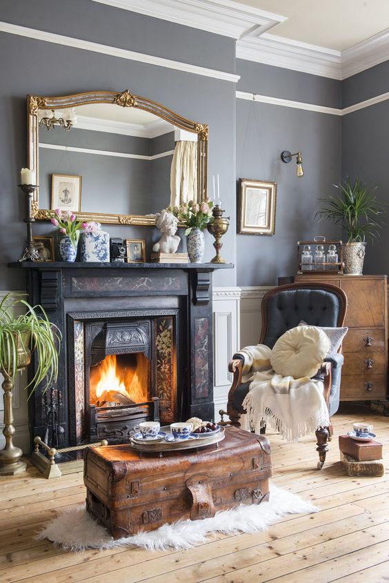 a vintage living roomw ith grey walls, a painted fireplace, a grey chair, a stained chest, greenery, a large mirror in a gilded frame