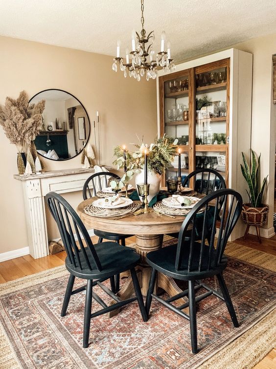 a vintage rustic dining space with a staiend round table, matching black chairs, a crystal chandelier, a glass buffet and a faux fireplace with a round mirror over the mantel