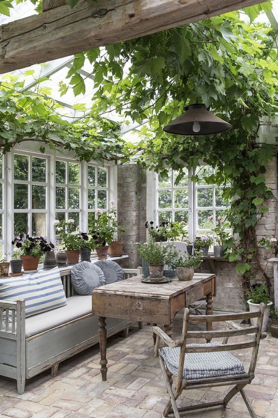 a vintage to shabby chic sunroom with neutral and stained furniture, potted greenery and blooms and vining greenery on the ceiling