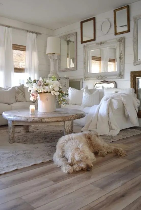 a white shabby chic living room with a gallery wall of mirrors, white furniture, a shabby table and lamps and blooms