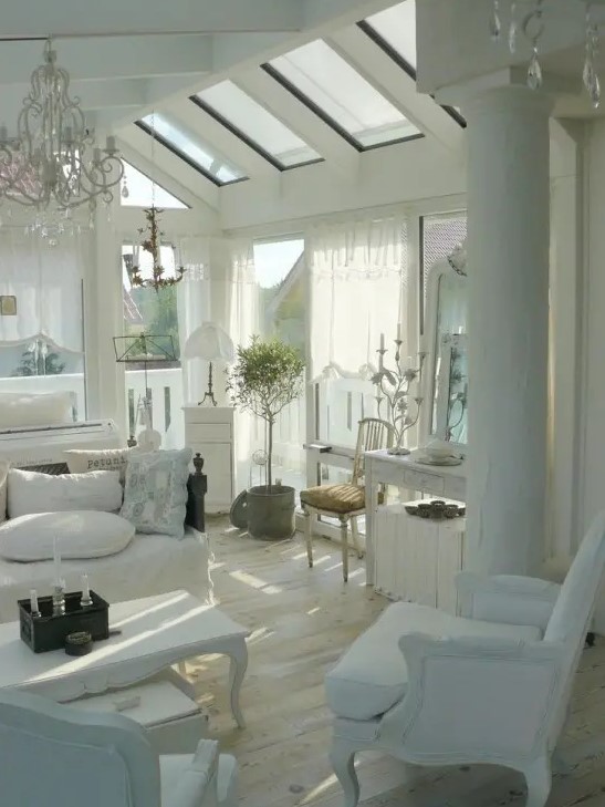 a white shabby chic living room with skylights, elegant white furniture, a crystal chandelier, a potted plant and greenery
