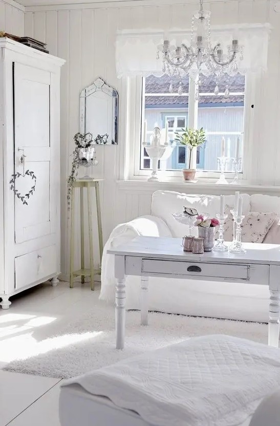 a white shabby chic living room with whitewashed furniture, a crystal chandelier, potted greenery and plants and floral pillows