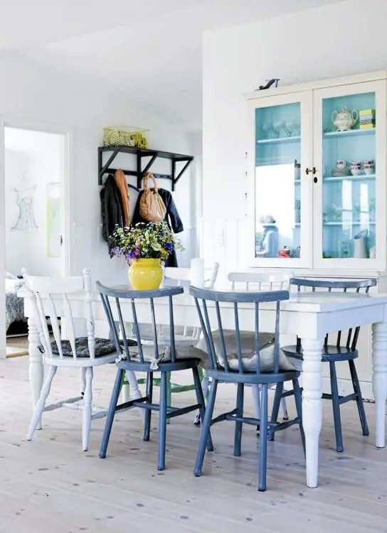 an airy Scandinavian dining space with a neutral cupboard with glass doors, a white vintage dining table, white and blue chairs and a bright vase with blooms