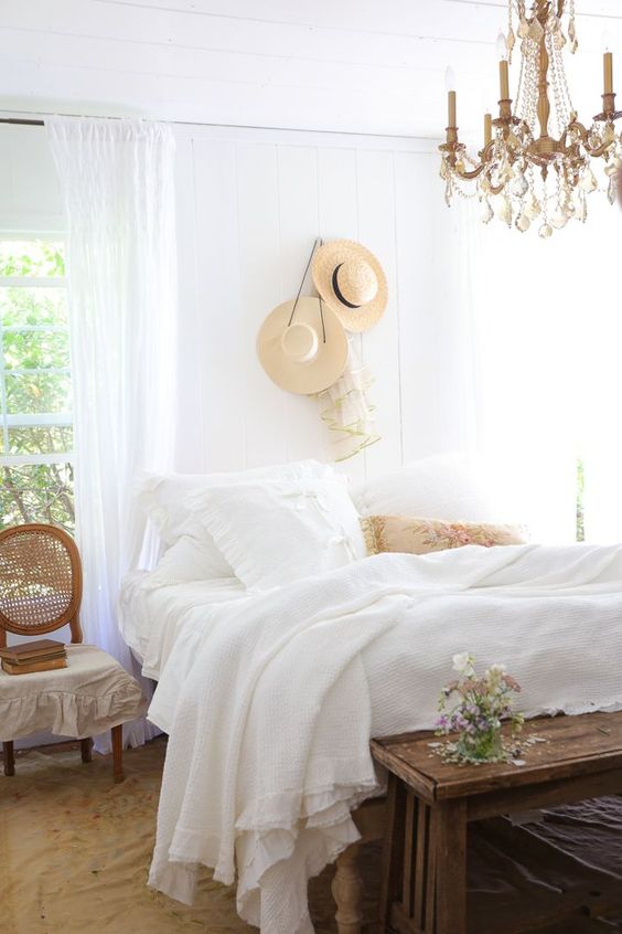 an airy and chic Provence bedroom with white walls and a ceiling, a bed with white bedding, a stained chair and a bench, a cool chandelier and straw hats