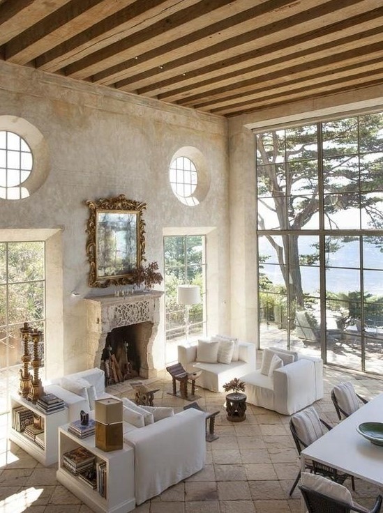 an airy and serene Provence living room with a fireplace, neutral furniture, wooden beams, bookshelves and a dining space