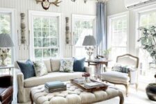 an airy vintage living room with chic vintage furniture, a larg eottoman, chic chandeliers, blue curtains and greenery