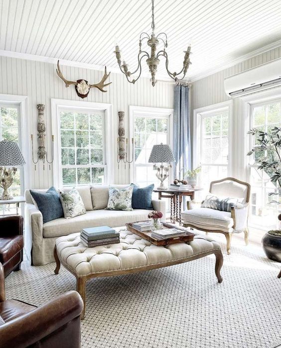 an airy vintage living room with chic vintage furniture, a larg eottoman, chic chandeliers, blue curtains and greenery