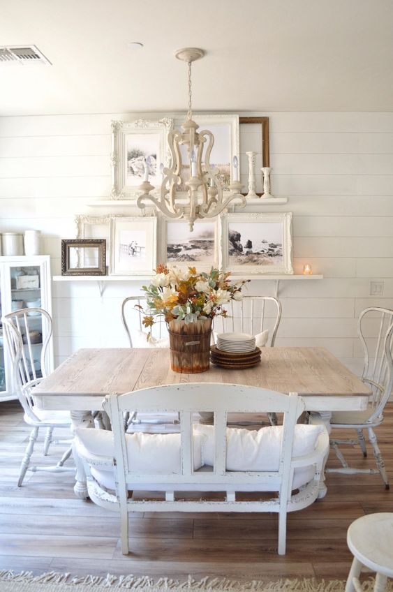 an all-white dining room with a gallery wall on ledges, a large dining table and chic vintage chairs and a loveseat, a white chandelier