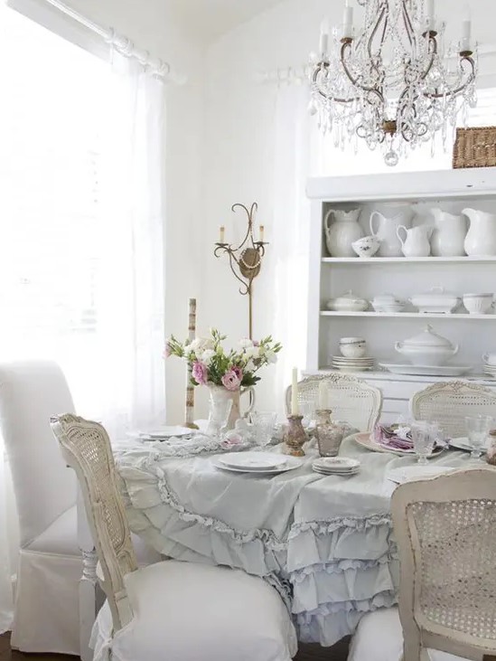 an elegant and neutral vintage dining room with a ruffle tablecloth, a crystal chandelier, shabby chic chairs and stylish porcelain