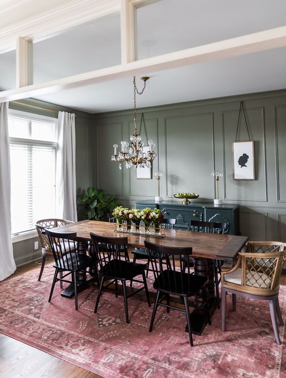 an elegant vintage-inspired dining room with olive green walls, a stained dining table, black wooden and stained rattan chairs, a chic chandelier