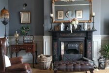 an elegant vintage living room with grey walls, a black fireplace, brown leather chairs and ottomans, a mini desk and artwork