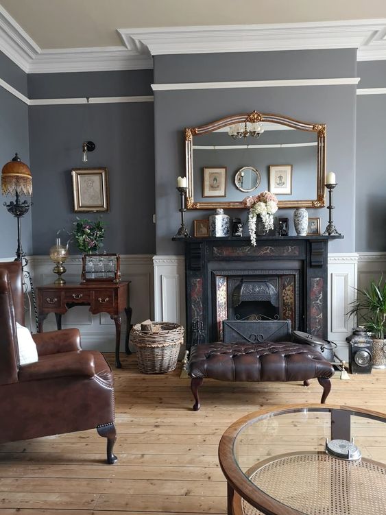 an elegant vintage living room with grey walls, a black fireplace, brown leather chairs and ottomans, a mini desk and artwork