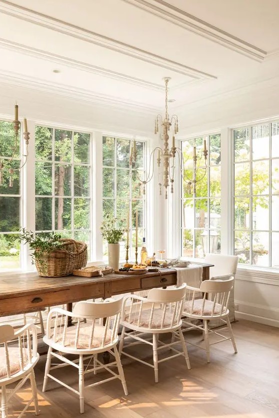 a French country dining room with a gallery of windows, a large stained table and vintage white chairs plus chandeliers