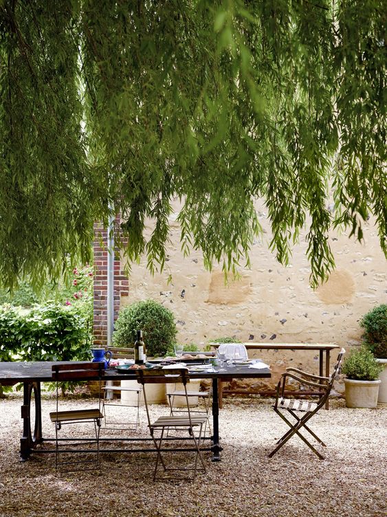 a Provence-inspired outdoor space with a stained table and metal and wood chairs, greenery and trees over the space