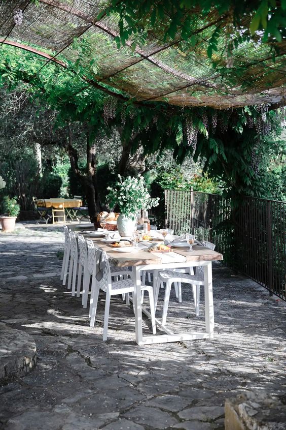 a Provence terrace with a roof with greenery, a metal and light-stained wood table and metal chairs, potted plants and blooms