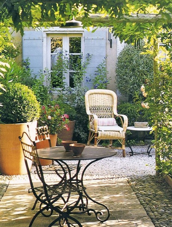 a Provence terrace with a woven chair, a metal table and some chairs, potted greenery and blooms and lots of sunlight
