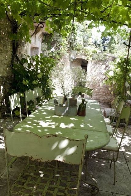 a Provence terrace with greenery, climbing plants and trees, a light green shabby chic dining set and pendant lamps