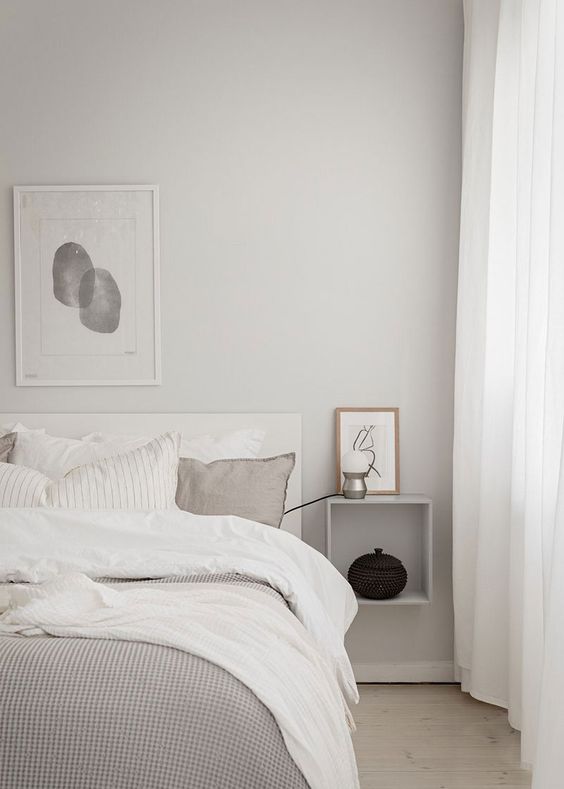 a beautiful minimalist bedroom with a monochromatic color scheme, light grey walls, a creamy bed with grey and white bedding, a wall-mounted nightstand and a floor lamp