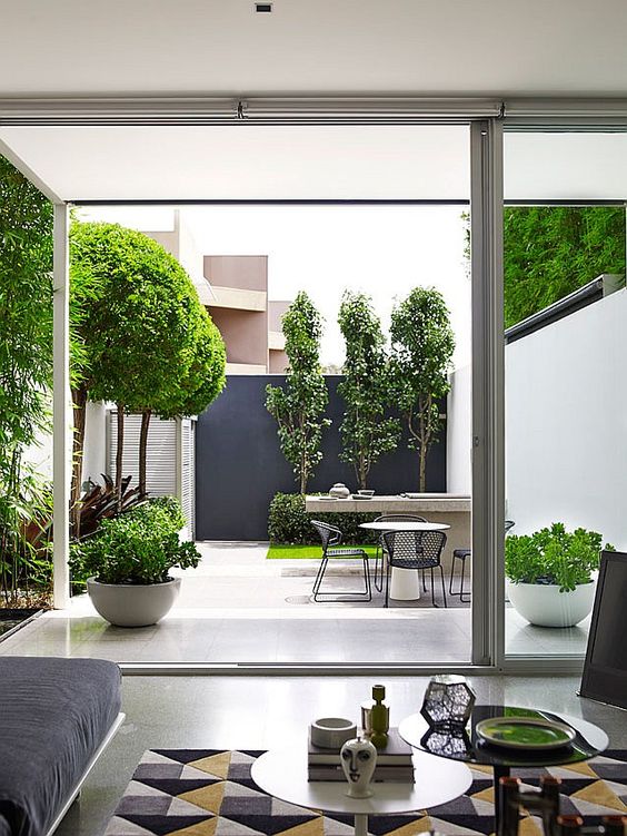 a catchy minimalist outdoor space with growing trees, grass and greenery, a stone table and metal chairs and a black accent wall