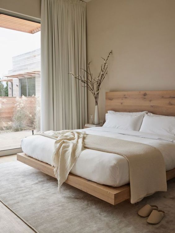 a dreamy neutral minimalist bedroom with tan walls, a stained bed with neutral bedding, light green curtains and blooming branches in a vase