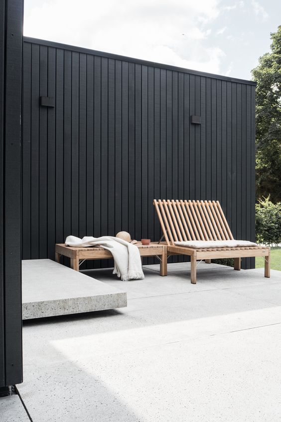 a fab minimalist terrace with black planked walls, a wooden lounger and a bench, a stone platform and a stone floor is a chic space