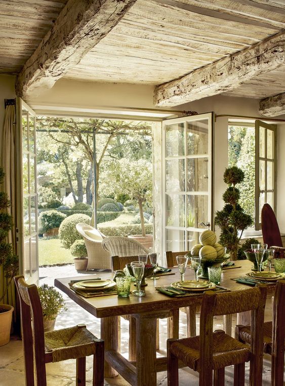 a lovely Provence dining room with large windows, a stained table and chairs, a whitewashed wooden ceiling with beams and lots of greenery